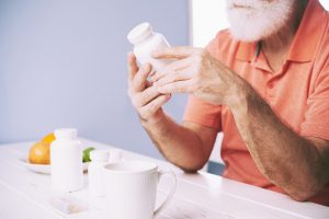 Hombre mayor con barba blanca y camisa naranja examinando atentamente un frasco de medicamento. En la mesa blanca frente a él hay otros frascos de pastillas, una taza y un pastillero, junto a un plato con frutas y verduras. El fondo es azul claro.