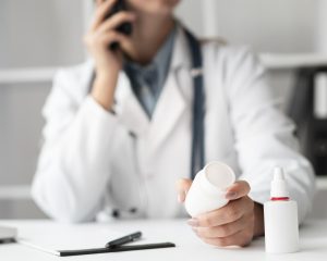 Una profesional de la salud sostiene un frasco de medicamento mientras atiende una llamada telefónica en su consultorio. La imagen refleja la importancia de la farmacovigilancia y el asesoramiento médico en el uso seguro de los fármacos.