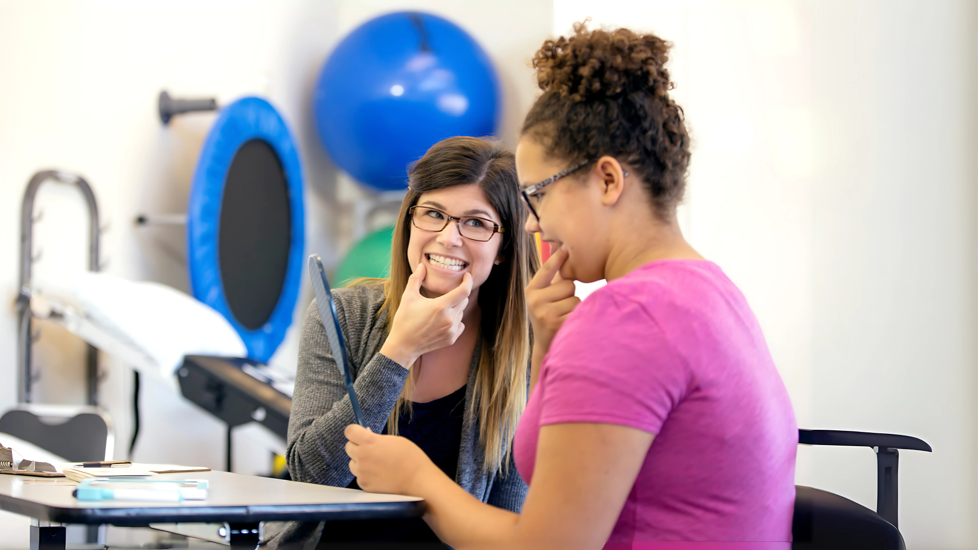 Dos mujeres realizan ejercicios faciales frente a un espejo como parte de una terapia miofuncional para tratar la apnea del sueño.