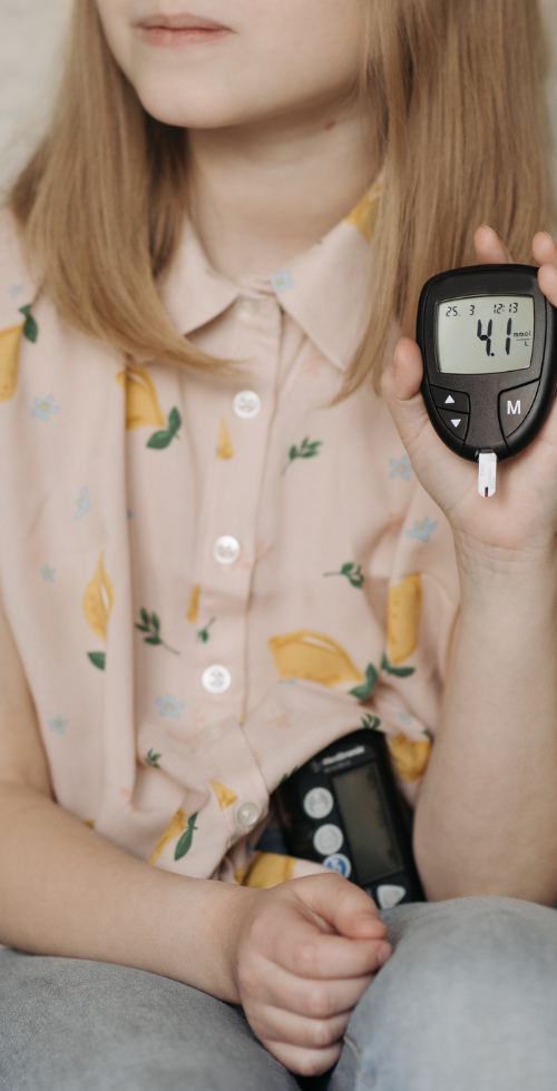 Niña sentada en un sofá sosteniendo un medidor de glucosa que muestra un valor bajo, con un dispositivo de control de diabetes apoyado en su regazo.